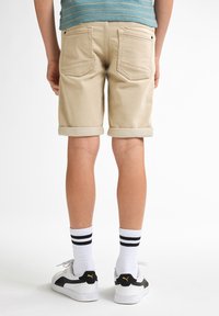 Pantalones cortos de algodón beige con puños enrollados, bolsillos traseros, usados con calcetines blancos con rayas negras y zapatillas en blanco y negro.