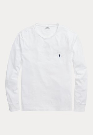 Un homme porte une chemise blanche à manches longues, un jean foncé, une casquette bleu marine et des baskets. Une chemise bleue à carreaux est nouée autour de sa taille.