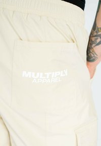 Beige cargo shorts med en elastisk talje og en baglomme med teksten "MULTIPLY APPAREL" i fed hvid skrift.