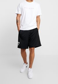 Camiseta blanca con el texto "BALL IS LIFE" en rosa, combinada con pantalones cortos deportivos negros con un diseño de palmeras blancas, y calzada con zapatillas blancas.