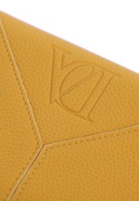 Superficie de cuero amarillo texturizado con letras entrelazadas en relieve y costuras visibles cosidas en un patrón geométrico.