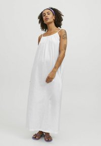 Robe longue en lin blanche avec des bretelles spaghetti fines, un col froncé, une coupe ample et une longueur jusqu'aux chevilles, associée à des tongs violettes.