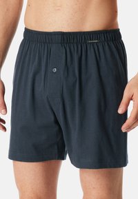 Sötétkék pamut boxeralsó, rugalmas derékrésszel, egyetlen elülső gombbal és laza szabással. Simma textúra, látható minták nélkül.