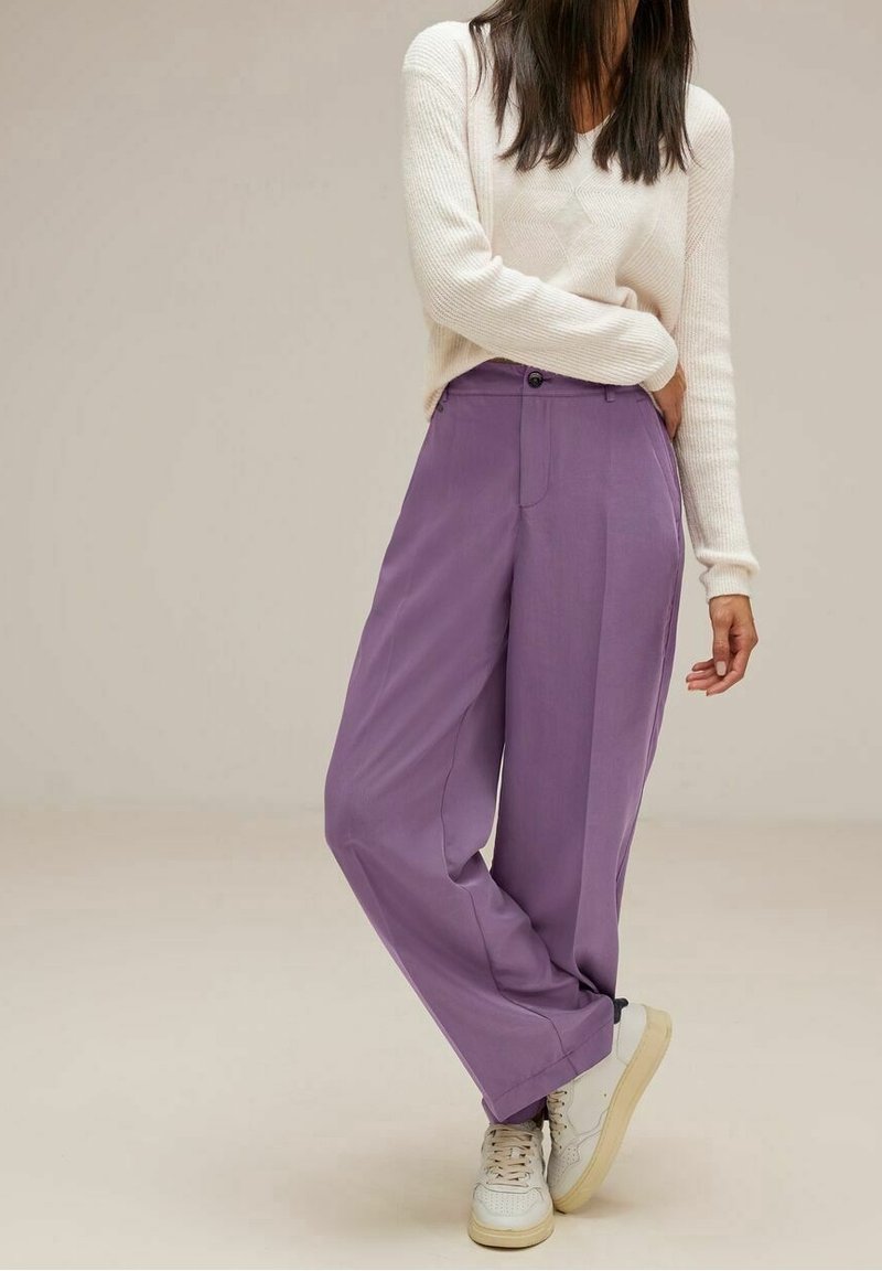 Lila, vida byxor med en slät textur; kombinerat med en stickad, krämfärgad tröja och vita sneakers. Enkelt design och rena linjer.