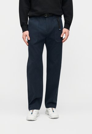 Mees, kes kannab tumesiniseid pükse suurte esiteks taskutega, musta vööd, musta pikkade varrukatega pluusi ning valgeid tossusid siniste ja punaste detailidega.