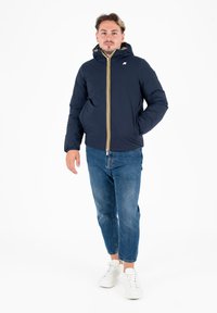 Giacca puffer blu navy con cappuccio, dotata di una zip anteriore gialla e bianca. Indossata sopra jeans blu e sneakers bianche. Design semplice.