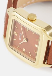 Montre bracelet en or carré avec un cadran marron, des chiffres romains et un bracelet en cuir marron. Présente un bracelet texturé et une couronne hexagonale.
