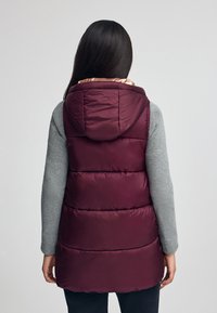 Burgundyröd pufferväst med huva, med horisontellt quiltade sektioner och ett lätt innerfoder. Bärs över en grå långärmad topp.