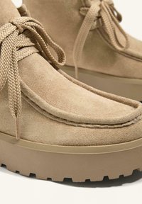 Béžové semišové členkové topánky s hrubými gumovými podrážkami, s textúrovanými šnúrkami a detailmi šitia; zaoblený tvar s jemným opätkom.