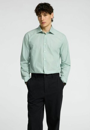 Camisa de rayas verde claro y blanca con mangas largas y cuello con botones, combinada con pantalones negros. Textura de tela suave.
