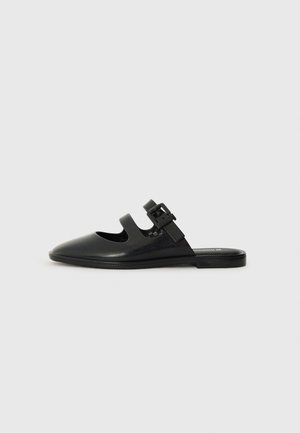 Black slip-on shoe made of glossy material, featuring cut-out design and adjustable strap with hardware accent. Flat sole with minimal tread.