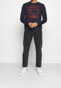 Felpa di cotone blu navy con logo grafico rosso "RAW" sul davanti, abbinata a jeans grigio scuro e scarpe da ginnastica bianche.