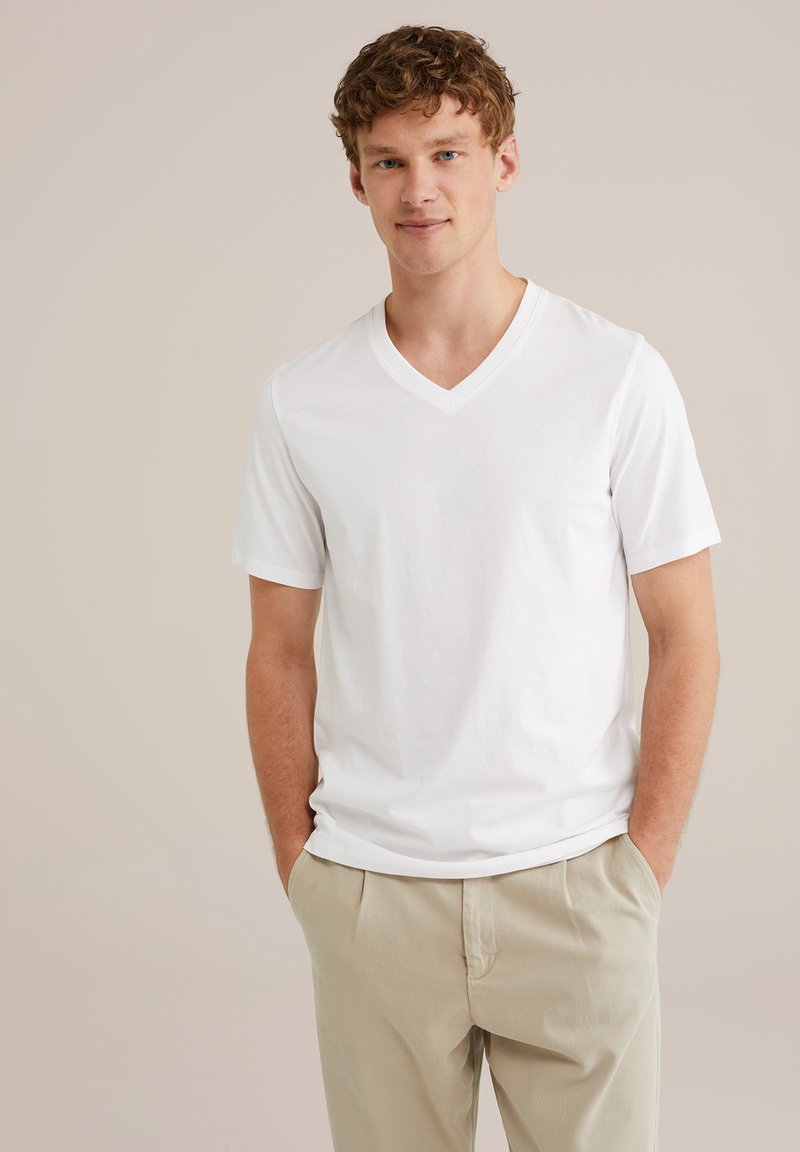Vit kortärmad V-ringad t-shirt i mjukt bomullstyg, med en avslappnad passform och subtila sömdetaljer. Kombinerad med ljusbeiga byxor.