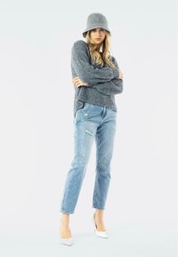 Maglione grigio a maglia, jeans chiari strappati, tacchi alti bianchi e un cappello a secchiello grigio in feltro. La modella è in piedi con le braccia incrociate, su uno sfondo neutro.