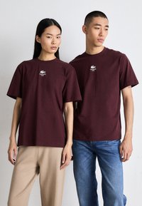 Bordové bavlněné trička s kulatým výstřihem a krátkými rukávy. Má vytištěné logo Lacoste na hrudi. Stylizováno s casual kalhotami a džíny.