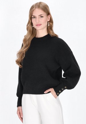 Zwarte gebreide trui met een relaxte pasvorm, geribbelde textuur en brede mouwen. Voorzien van knoopaccenten bij de manchetten. Gecombineerd met witte broek.