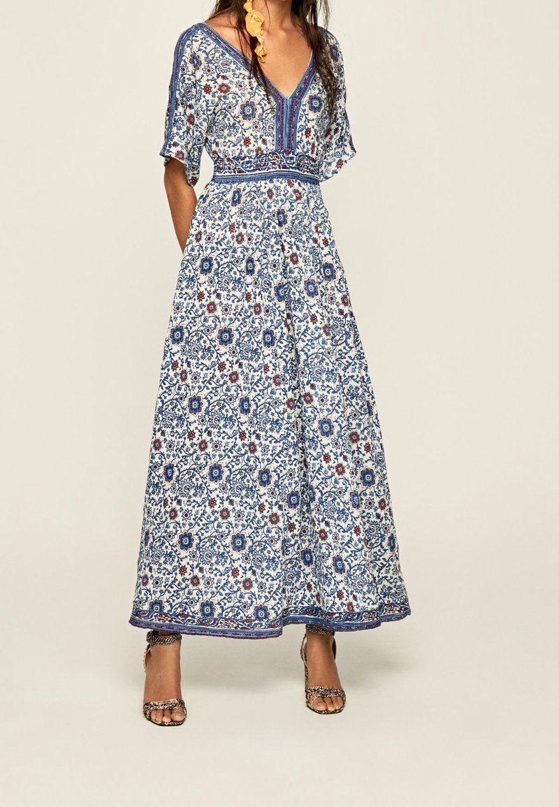 Femme portant une robe longue blanche avec des motifs floraux bleus et rouges, col en V, manches courtes, et associée à des sandales à talons hauts.