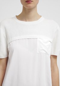 T-shirt bianco con un pannello superiore testurizzato e tessuto liscio nella parte inferiore. Include una piccola tasca, maniche corte e un collo rotondo.