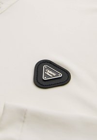 Tessuto beige con una superficie liscia, caratterizzato da una patch in pelle nera a forma di triangolo con la parola "OMBRE" in bianco e dettagli argentati.