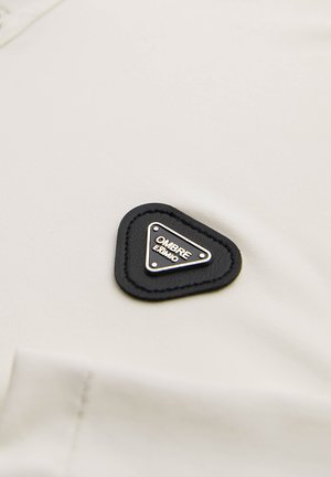 Beige stof med en glat tekstur, der har en sort trekantet læderpatch med "OMBRE" i hvid tekst og sølvfarvede detaljer.