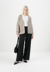 Beige huňatý kardigan s otevřeným předním dílem, nošený přes bílý top. Černé široké kalhoty a černé špičaté lodičky. Model drží černou tašku.