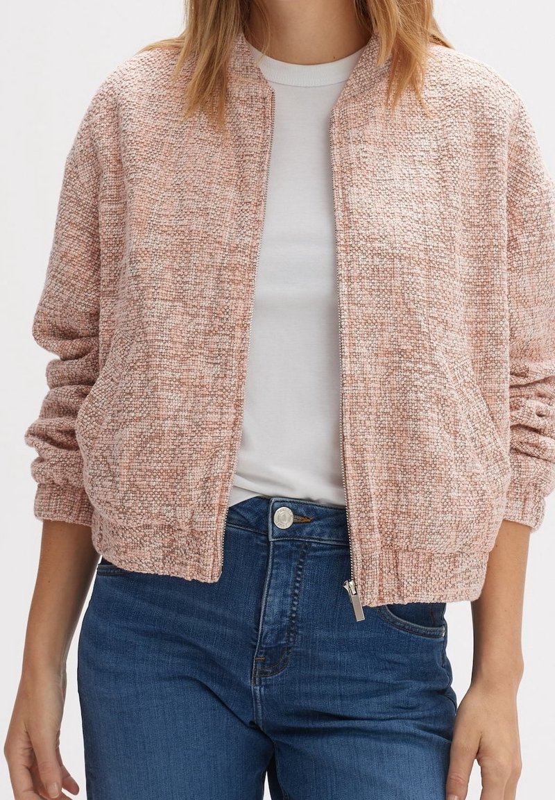 Femme portant une veste zippée rose clair texturée sur un t-shirt blanc et un jean bleu, debout les bras détendus le long du corps.