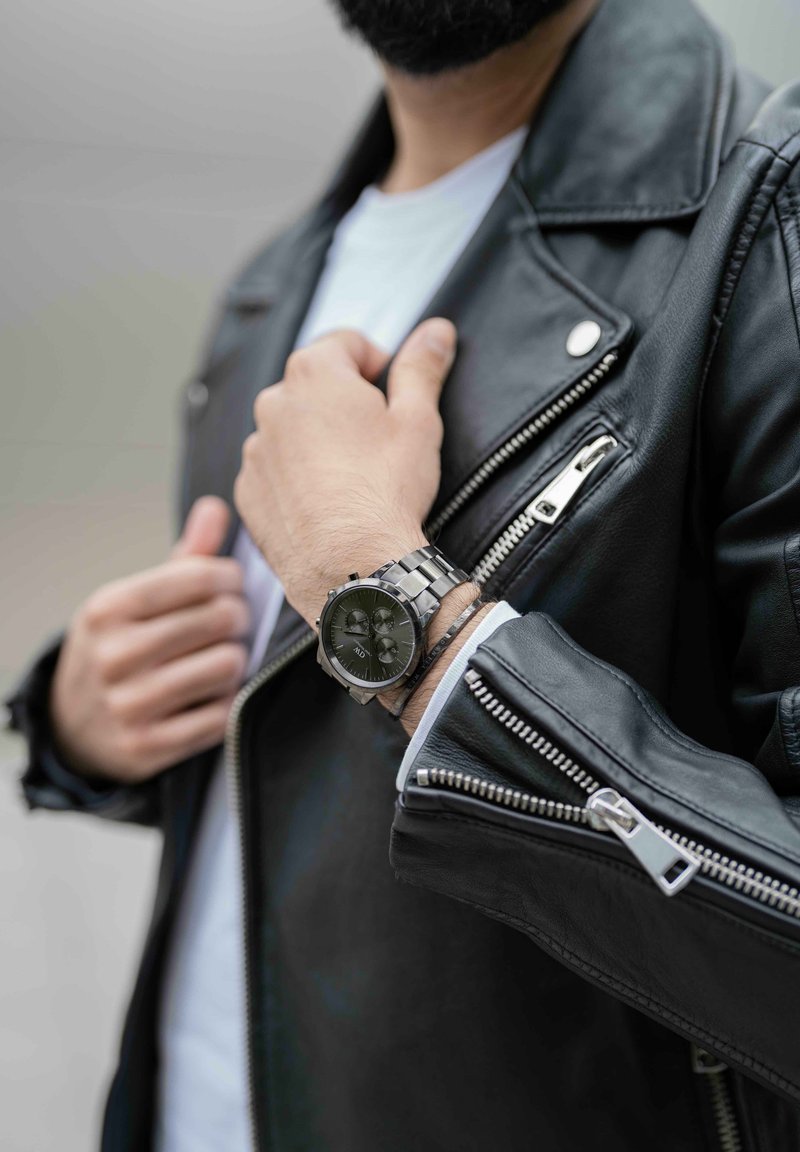 Montre-bracelet en métal argenté avec un cadran noir et des fonctionnalités de chronomètre, portée sur une main dans une veste en cuir noir avec des zips argentés.