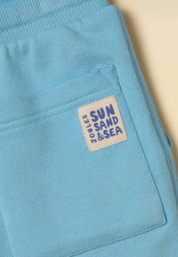 Svetlomodrá látka s prešitým štvorecovým vreckom, na ktorom je béžová etiketa s modrým nápisom „Joules Sun Sand & Sea“.