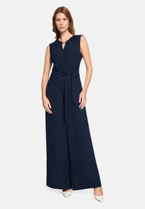 Combinaison sans manches bleu marine avec un col plissé, une ceinture à nouer à la taille et des jambes larges, accompagnée de talons beige.