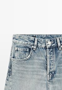 Lichtblauwe spijkerbroek met zichtbare tailleband, riemlussen, knoop en voorzak met muntenzak op een witte achtergrond.