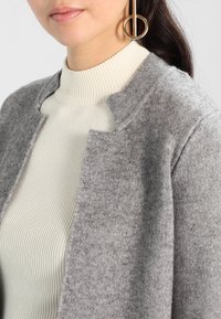 Kobieta w kremowym swetrze z golfem w prążki, szarym wełnianym kardiganie zapinanym na przód oraz dużych złotych geometrycznych kolczykach w kształcie koła.