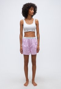 Γυναίκα με σγουρά μαλλιά, που φοράει λευκό crop top και ροζ ριγέ σορτς, στέκεται ξυπόλυτη μπροστά σε απλό φόντο.