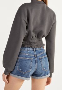 Grau beschnittenes Sweatshirt mit gerafftem Saum, langen Ärmeln und geripptem Kragen, kombiniert mit hellblauen, hochsitztenden Jeans-Shorts.