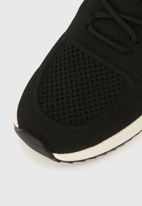 Juodi megzti sportbačiai su tekstūruotu viršumi, perforuotais elementais ir balta vidpadžio dalimi. Pado apačioje yra tekstūruota sukibimo raštas.