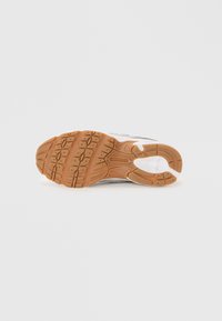 Chaussure de sport avec une semelle extérieure en caoutchouc arbore un motif de crampons marron, une semelle intermédiaire blanche et un dessus gris. Design minimaliste avec une forme arrondie.
