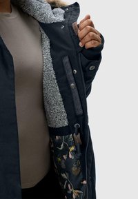 Person som håller upp en svart vinterjacka med grå fleecefoder och blommönstrad innerficka.