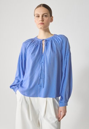 Blusa a righe blu con una patta bottonata, scollo arricciato e maniche lunghe ampie. Tessuto morbido, vestibilità comoda, abbinata a pantaloni bianchi.