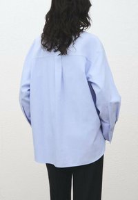 Camicia oversize di colore azzurro chiaro realizzata in tessuto morbido, con una piega sul retro e polsini con bottoni, abbinata a pantaloni neri.
