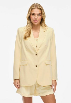 Light yellow blazer with a notched lapel, two front pockets, and brown buttons. Worn over a matching top and shorts, featuring smooth fabric.