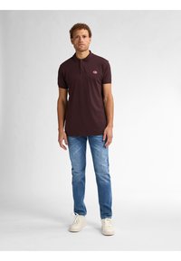 Ciemnobrązowa koszulka polo z trzema guzikami oraz małym logo na klatce piersiowej, w zestawieniu z jasnoniebieskimi dżinsami i białymi sneakersami.