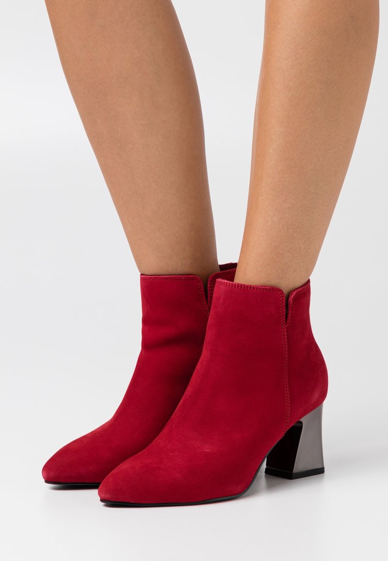 Bottines rouges en daim avec bouts pointus et talons blocs portées sur jambes nues sur un fond clair uni.
