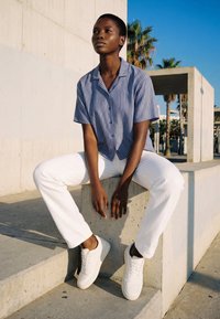 Jong persoon met kort haar, die een blauw gestreept shirt, witte broek en witte sneakers draagt, zit op betonnen trappen buiten.