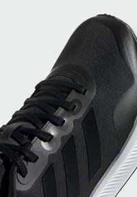 Zapatilla deportiva negra con parte superior de tela texturizada, acentos sintéticos, cordones planos y una suela de goma blanca con patrones de tracción.