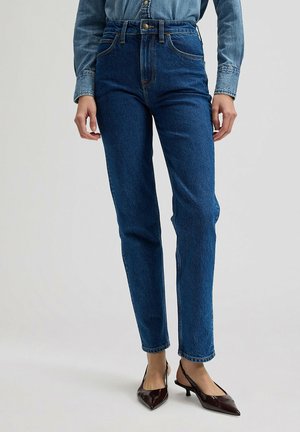 Kobieta ubrana w ciemnoniebieskie jeansy z wysokim stanem, wpuszczoną jeansową koszulę oraz bordowe szpilki z odkrytą piętą, stojąca na jednolitym tle.