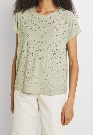 Mujer que lleva una blusa de manga corta verde claro con bordado calado y pantalones blancos, de pie sobre un fondo liso.