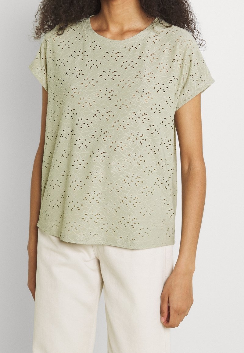 Femme portant un chemisier vert clair à manches courtes avec broderie ajourée et pantalon blanc, debout devant un fond uni.
