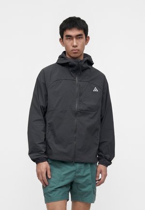 Jeune homme portant une veste à capuche noire avec fermeture éclair, un logo triangulaire et un short bleu sarcelle, debout devant un fond blanc uni.