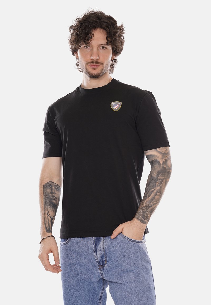 Uomo con capelli ricci indossa una maglietta nera con piccolo logo sul petto e jeans azzurri, mostrando tatuaggi a manica lunga su entrambe le braccia.