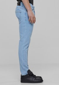 Svijetloplave traperice slim fit kroja, s tradicionalnim dizajnom od pet džepova i crnom oznakom straga. Nosive s crnim tenisicama.