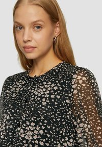 Schwarze Bluse mit floralem Muster in Pink und Weiß. Durchsichtige Puffärmel und Knopfverschluss am Hals. Weicher Stoff und elegantes Design.